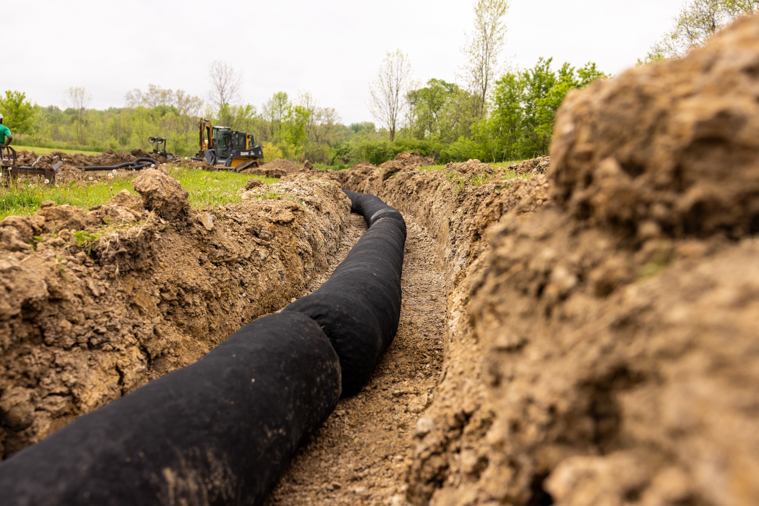 Excavator near trench with drainage pipe installation.