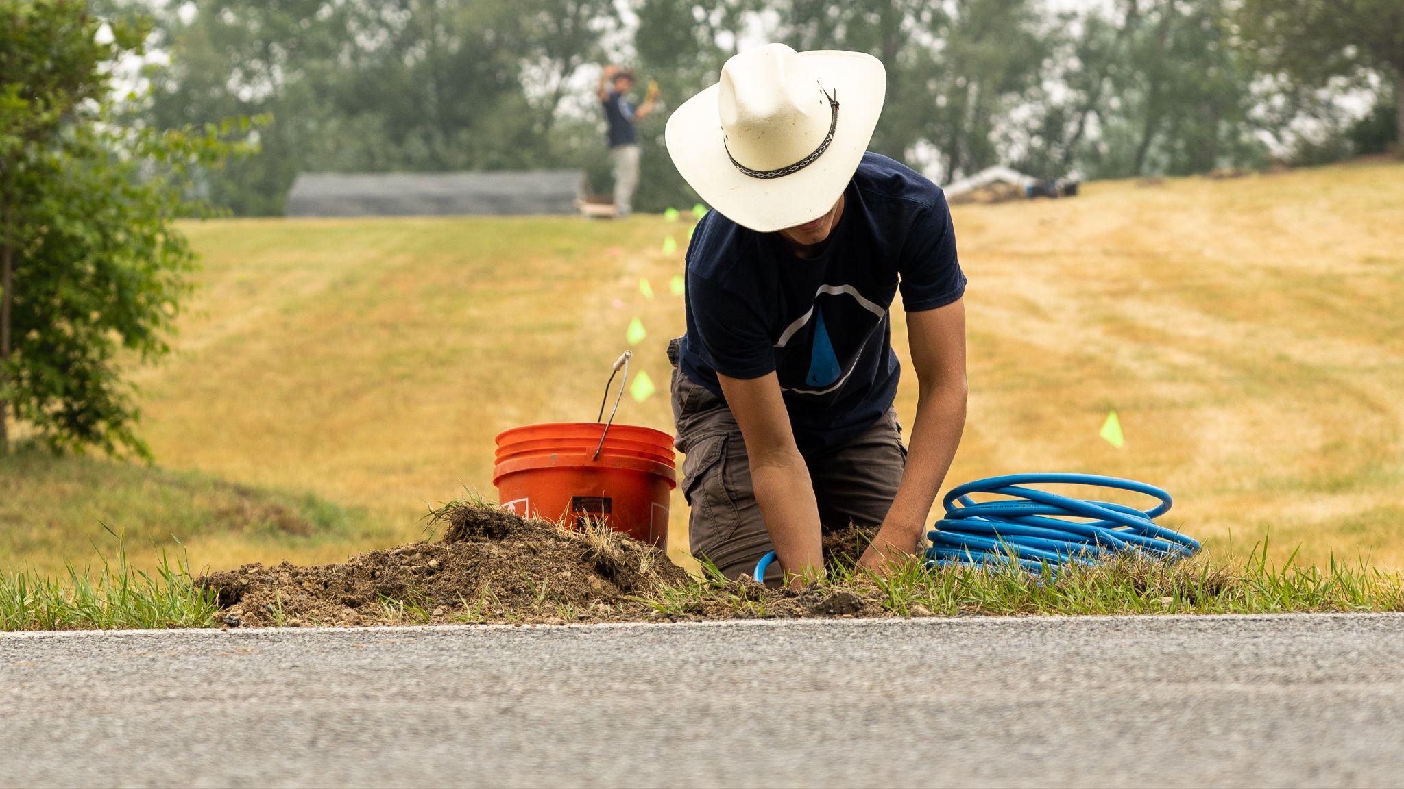 Worker installing irrigation line in dry soil with bucket and tubing nearby