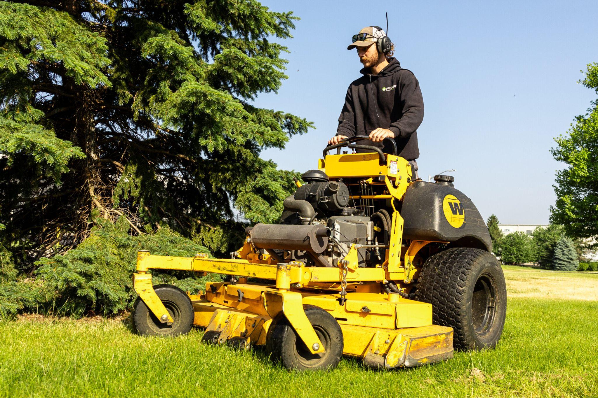 Worker riding a yellow commercial mower cutting grass near trees
